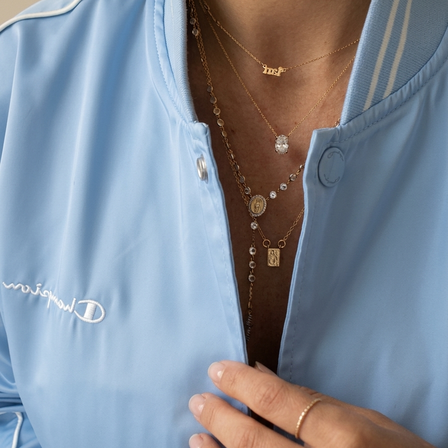 Woman wearing layered fine jewelry necklaces including a 14K gold Virgen del Carmen and Sacred Heart scapular necklace, miraculous medal rosary chain, oval diamond pendant, and Jesus name necklace styled with a light blue jacket.