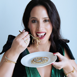 Woman eating pasta with a smile, holding a plate of food.