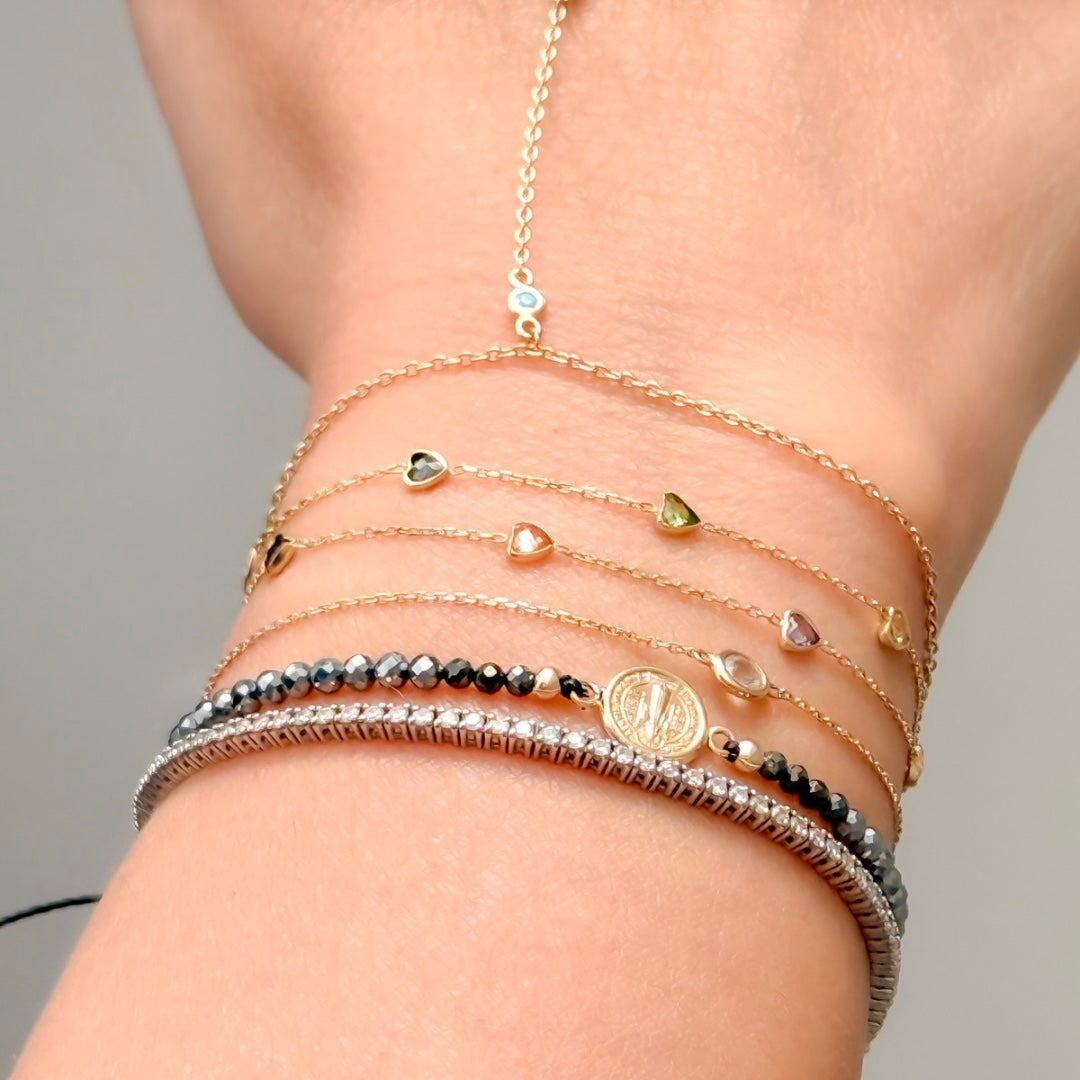 Layered bracelets on wrist including 14K gold gemstone chain bracelets, terahertz bead bracelet and 14K solid gold Saint Benedict medal bracelet.