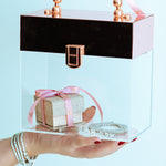 Clear box with a black lid and rose gold handle, containing a small gift wrapped in pink ribbon, held by a hand against a light blue background.