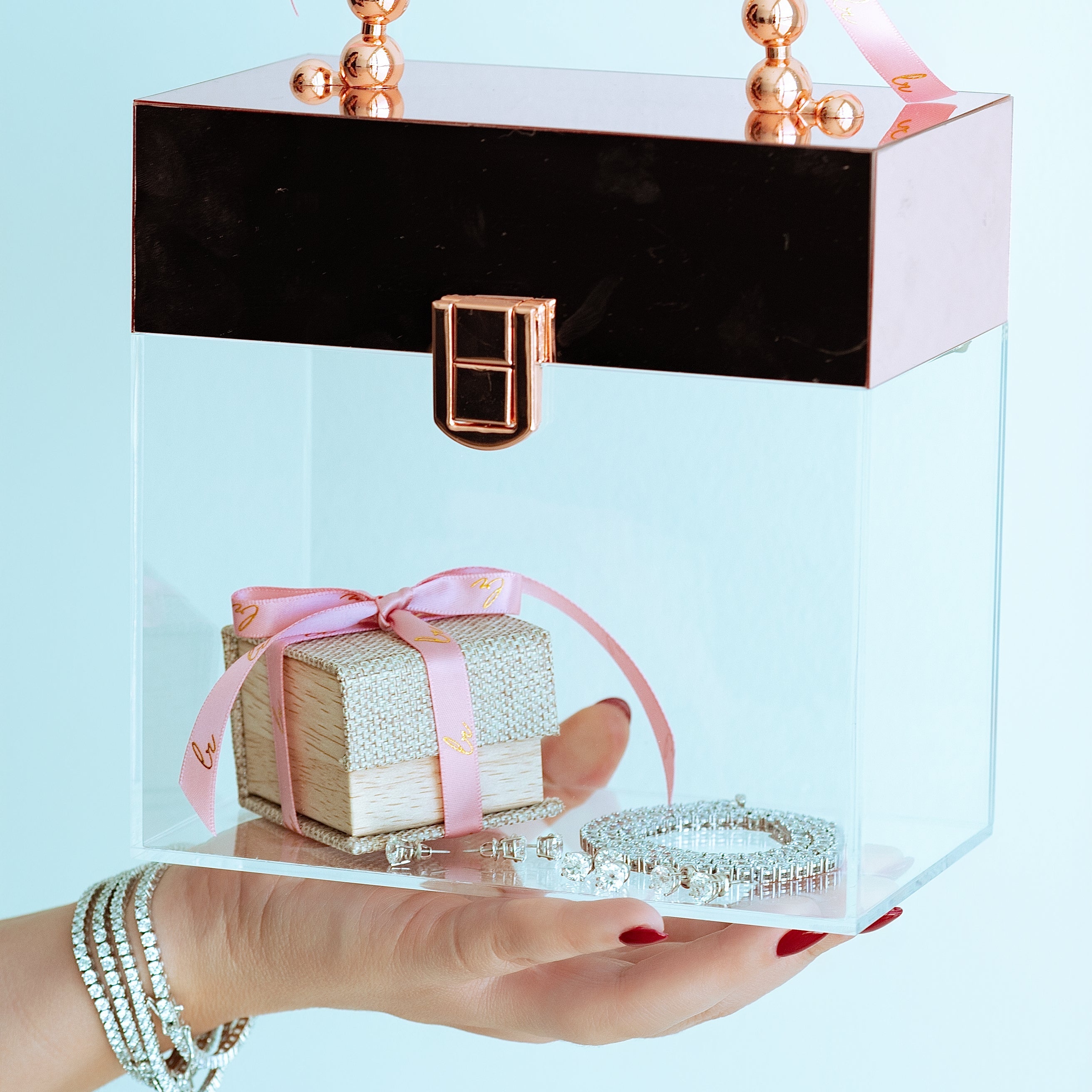 Clear box with a black lid and rose gold handle, containing a small gift wrapped in pink ribbon, held by a hand against a light blue background.