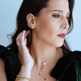 Woman wearing gold jewelry including earrings, necklace, and bracelet against a light blue background