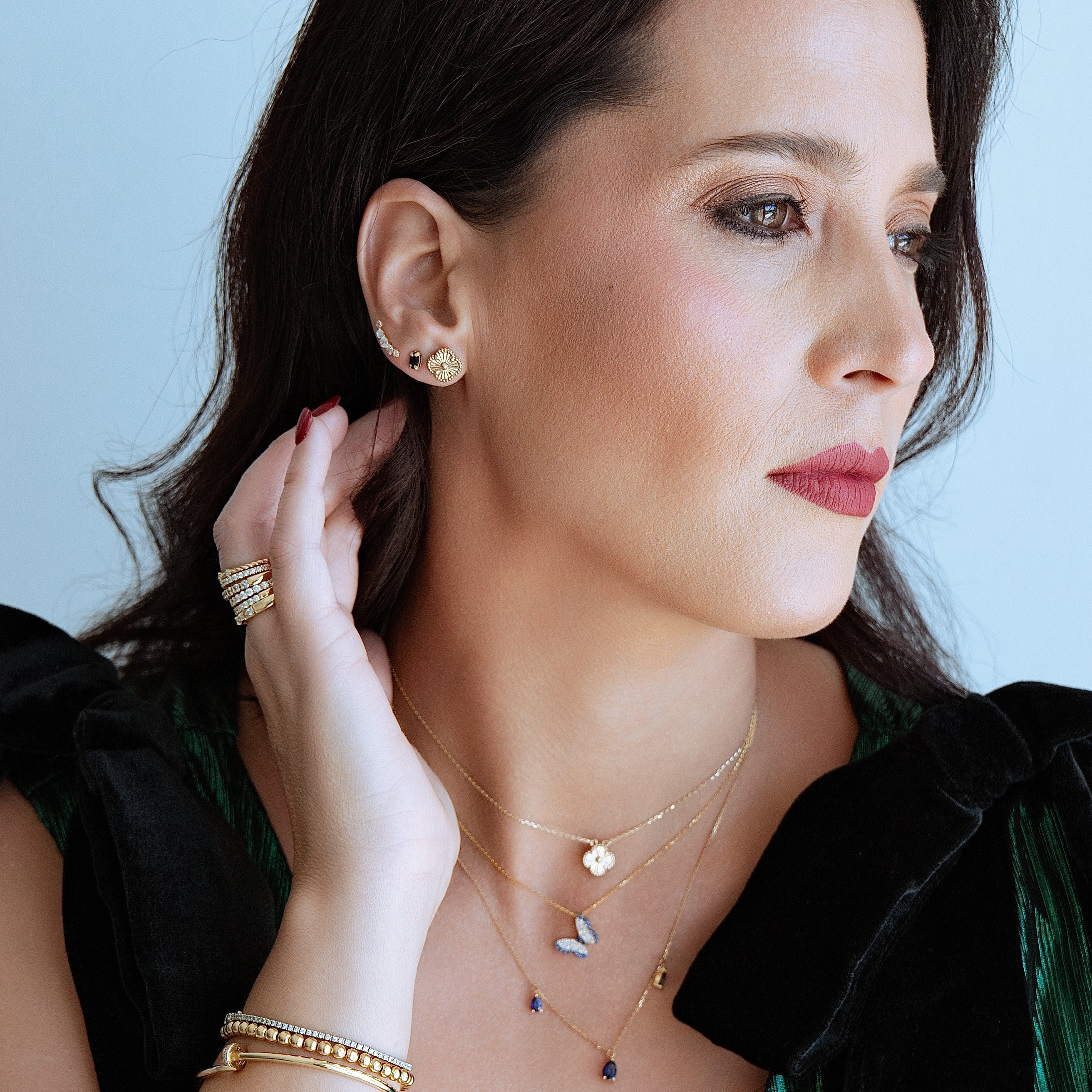 Woman wearing gold jewelry including earrings, necklace, and bracelet against a light blue background