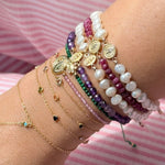 Close-up of wrist stacked with 14K gold bracelets featuring freshwater baroque pearls, amethyst gemstones, ruby beads, green stones, Miraculous Medals, Saint Benedict charms, and delicate gold chain bracelets over pink striped fabric