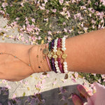 Wrist stacked with 14K gold faith bracelets featuring freshwater pearls, amethyst, ruby gemstones, Miraculous Medals, and delicate gold chains photographed outdoors with pink flowers on the ground