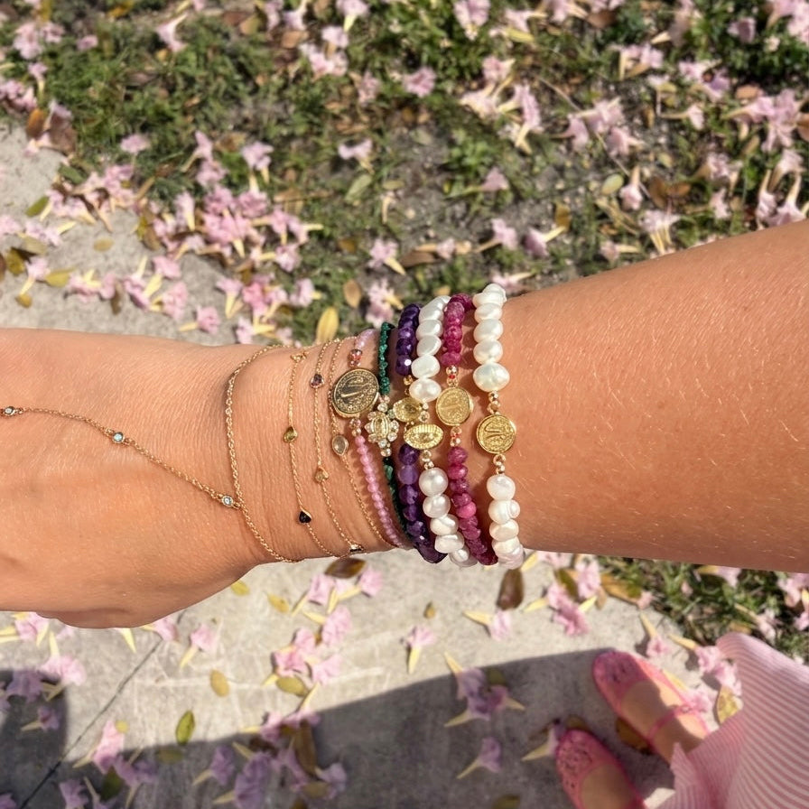 Wrist stacked with 14K gold faith bracelets featuring freshwater pearls, amethyst, ruby gemstones, Miraculous Medals, and delicate gold chains photographed outdoors with pink flowers on the ground