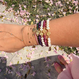 Wrist stacked with 14K gold faith bracelets featuring freshwater pearls, amethyst, ruby gemstones, Miraculous Medals, and delicate gold chains photographed outdoors with pink flowers on the ground