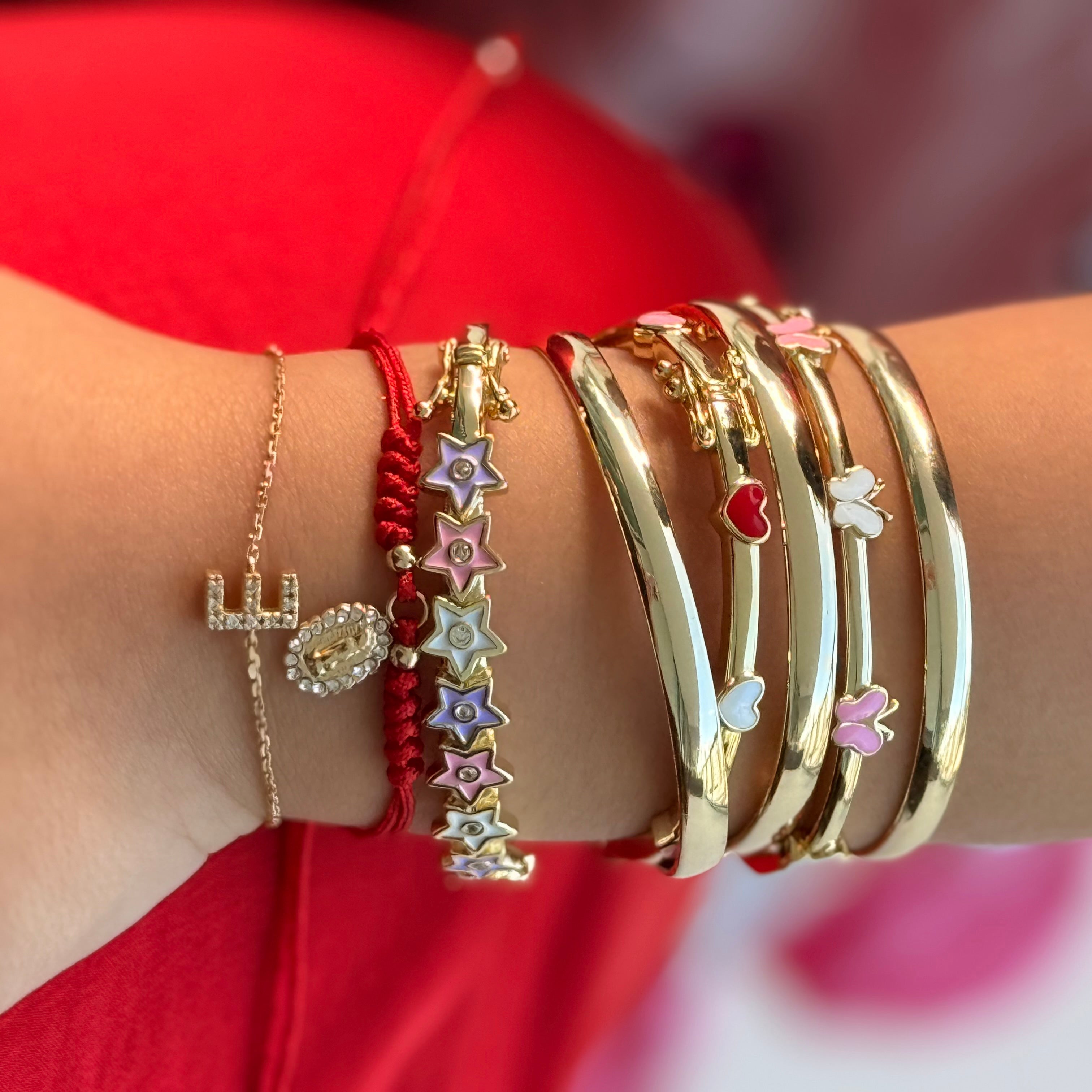 Close-up lifestyle photo of a wrist stacked with 14K gold bracelets, colorful enamel bangles, and a red adjustable string bracelet featuring a CZ Guadalupe Virgin charm, styled with a diamond initial bracelet and pink outfit details.