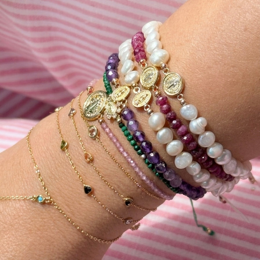 Wrist stacked with 14K gold faith bracelets featuring freshwater pearls, amethyst, ruby gemstones, Miraculous Medals, and delicate gold chains photographed outdoors with pink flowers on the ground