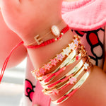 Lifestyle wrist stack featuring a 14K gold diamond initial bracelet, red adjustable Guadalupe Virgin string bracelet, pastel enamel star bracelet, and polished gold bangles with heart and butterfly accents.