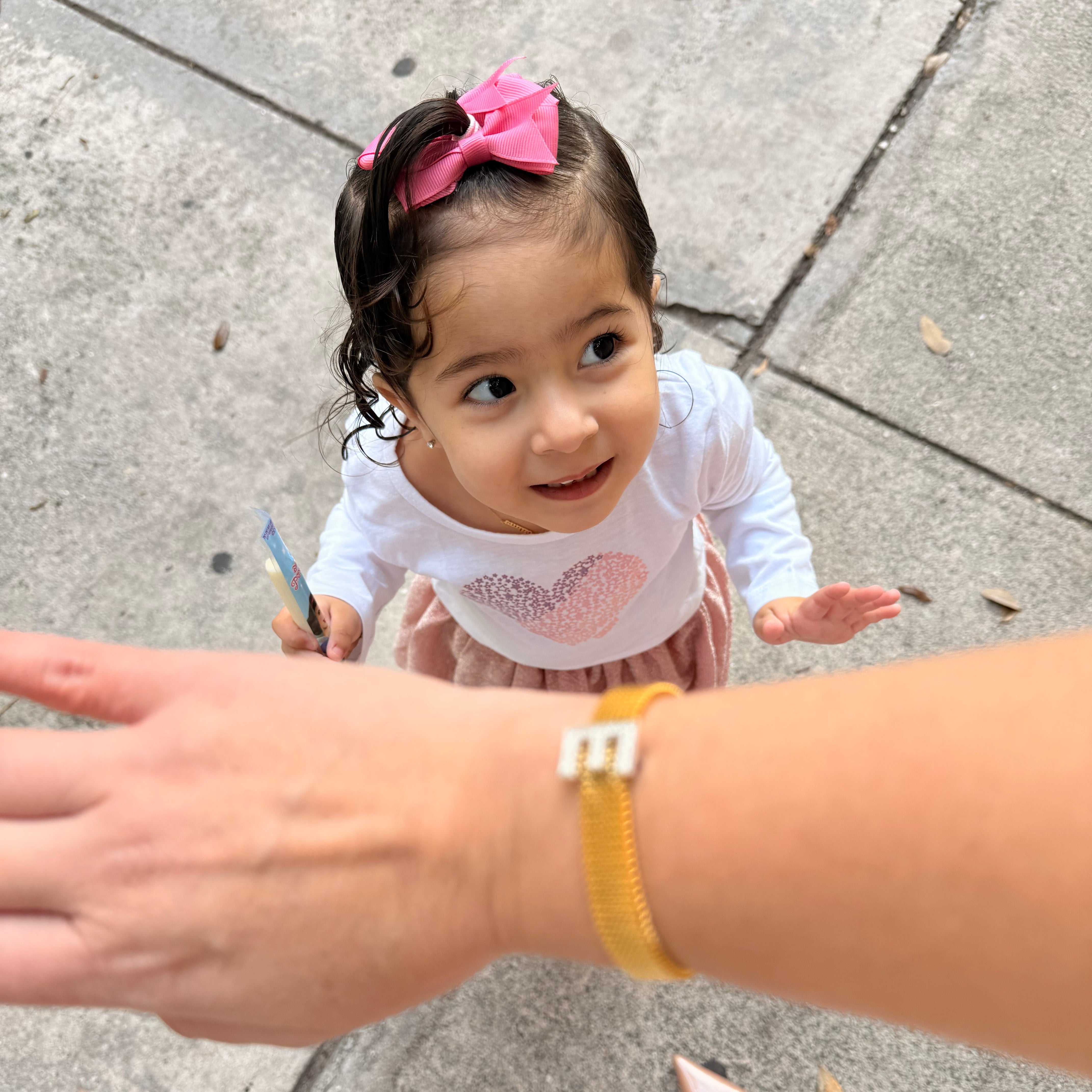 Child with a pink bow and white outfit on a sidewalk, with a hand reaching out.