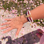 Wrist stacked with 14K gold faith bracelets featuring freshwater pearls, amethyst, ruby gemstones, Miraculous Medals, and delicate gold chains photographed outdoors with pink flowers on the ground