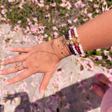 Wrist stacked with 14K gold faith bracelets featuring freshwater pearls, amethyst, ruby gemstones, Miraculous Medals, and delicate gold chains photographed outdoors with pink flowers on the ground