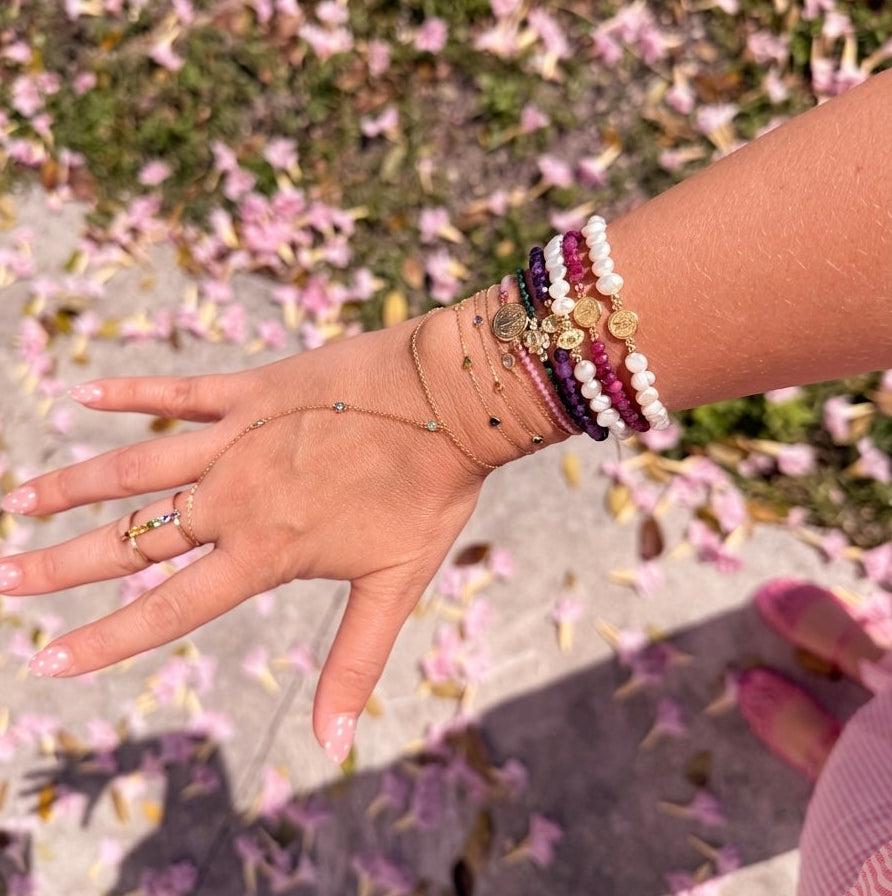 Wrist stacked with 14K gold faith bracelets featuring freshwater pearls, amethyst, ruby gemstones, Miraculous Medals, and delicate gold chains photographed outdoors with pink flowers on the ground