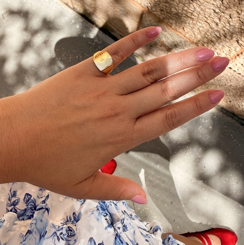 Close-up lifestyle photo of a hand wearing a 14K solid yellow gold square signet ring with a polished flat top, styled with soft pink nails and a blue floral dress.