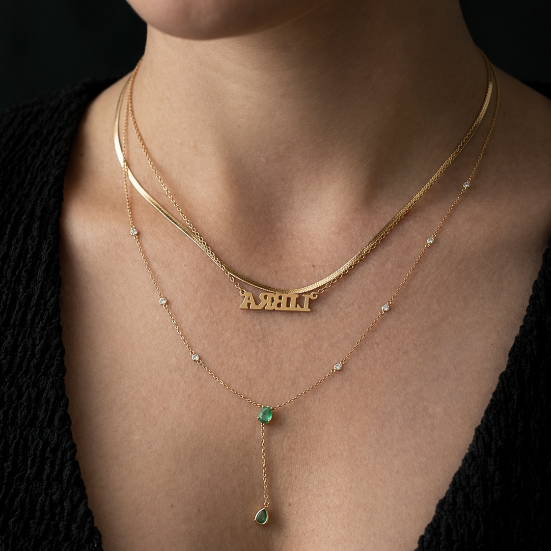 Close-up of a person wearing a gold necklace with a name pendant and a green gemstone on a black background.