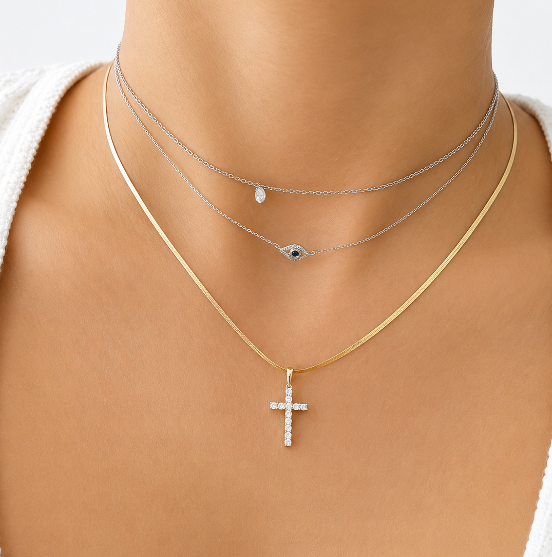 14K white gold diamond cross pendant necklace layered with a white gold evil eye necklace and a white gold bezel solitaire choker on model, minimalist fine jewelry stack.