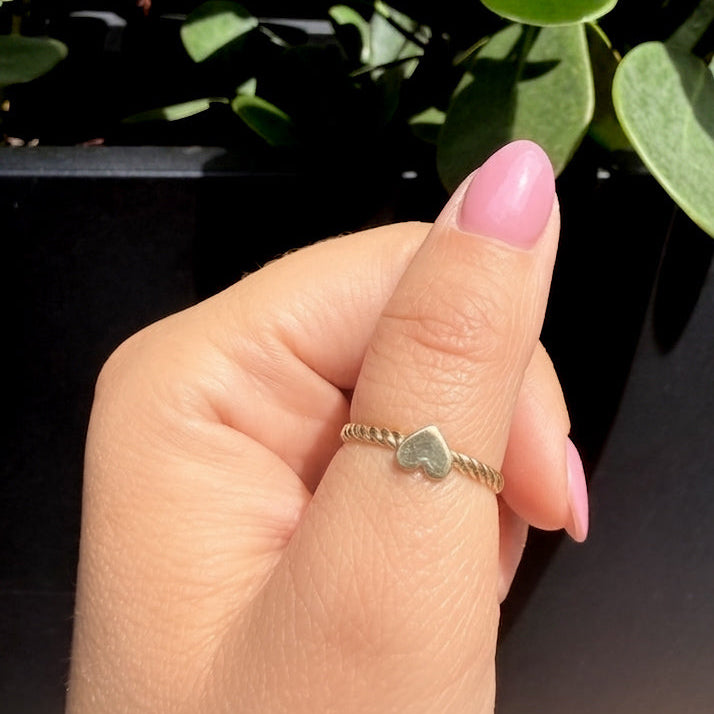 Close-up of a hand wearing a 14K solid yellow gold heart ring with twisted rope band, polished heart detail shining under natural sunlight with green plants in the background