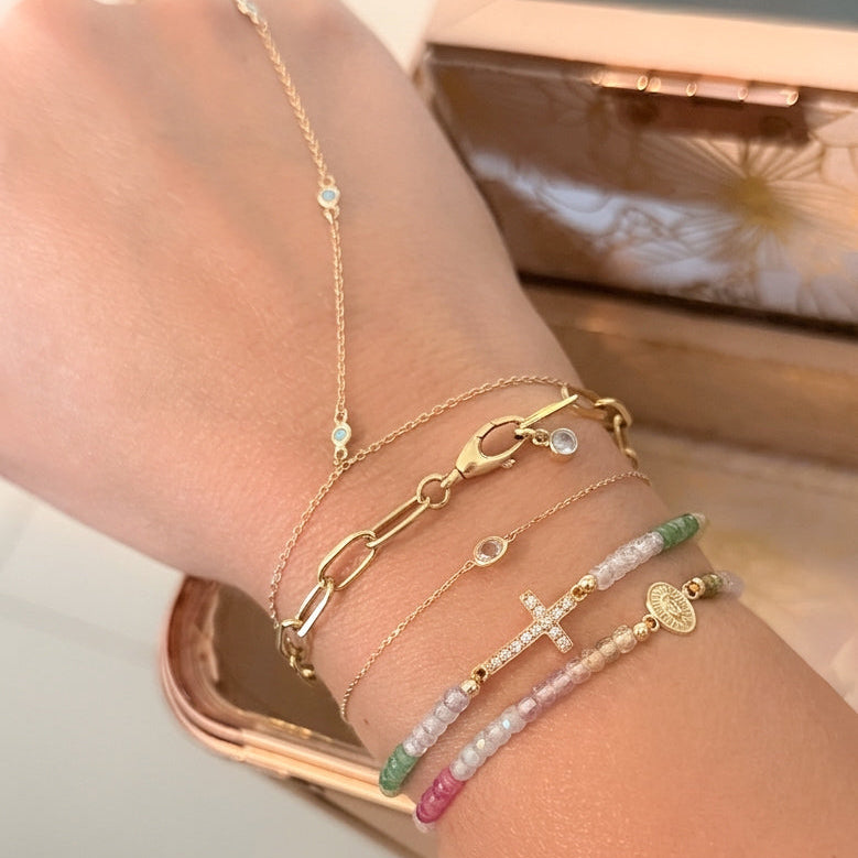 14K yellow gold layered bracelet stack on wrist featuring evil eye hand chain bracelet, paperclip chain bracelet, bezel diamond bracelet, diamond cross gemstone bracelet, Virgin Mary medallion bracelet, and multicolor gemstone beaded bracelet styled in soft natural light