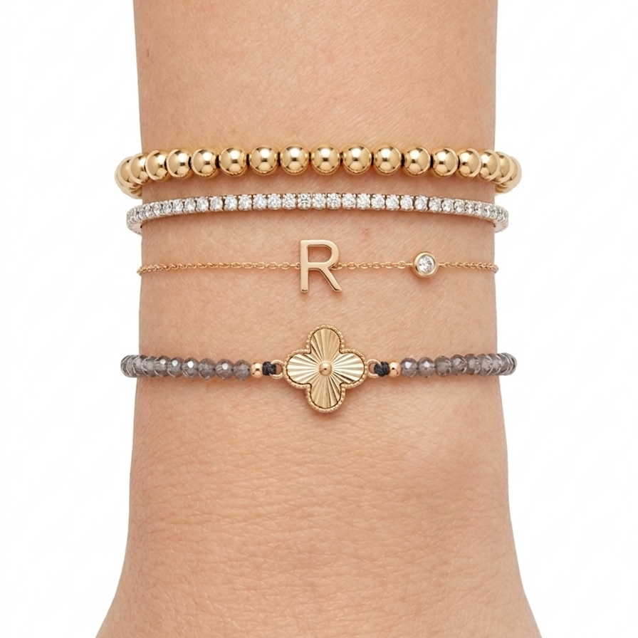 Luxury wrist stack with 14K gold bead bracelet, diamond tennis bracelet, initial diamond bracelet, and gray gemstone clover bracelet layered together.