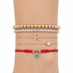 14K solid yellow gold evil eye red string bracelet styled on wrist with gold bead bracelet, diamond tennis bracelet, and initial R necklace bracelet stack on white background