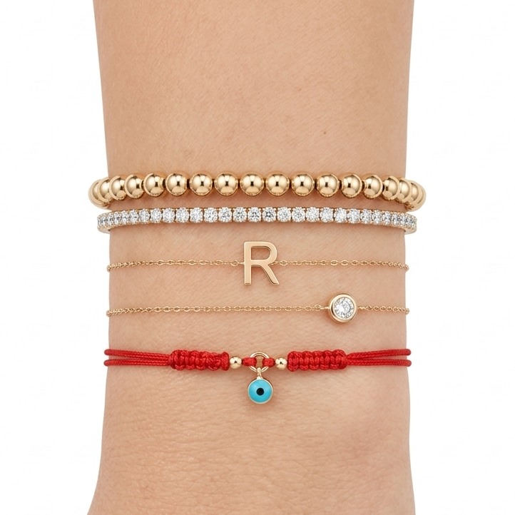 14K solid yellow gold evil eye red string bracelet styled on wrist with gold bead bracelet, diamond tennis bracelet, and initial R necklace bracelet stack on white background