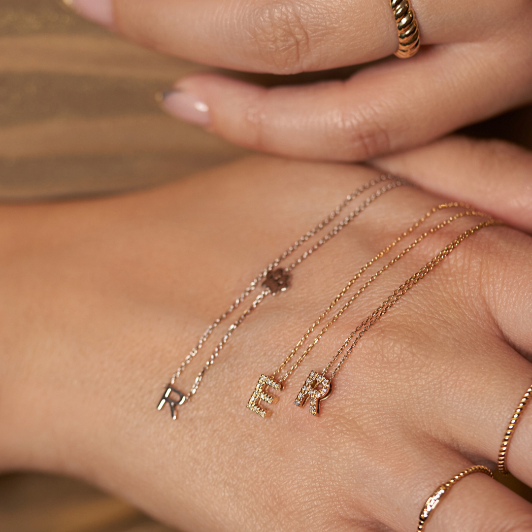 Initial necklaces displayed across a hand, including a white gold R and paw print necklace beside gold E and R necklaces.