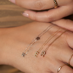 Initial necklaces displayed across a hand, including a white gold R and paw print necklace beside gold E and R necklaces.